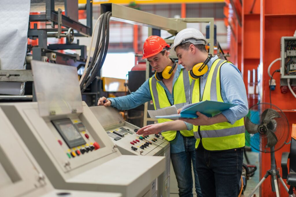 Männliche Techniker tragen eine Sicherheitsweste mit Helm, während sie in der Fabrik Maschinen überprüfen und reparieren. 