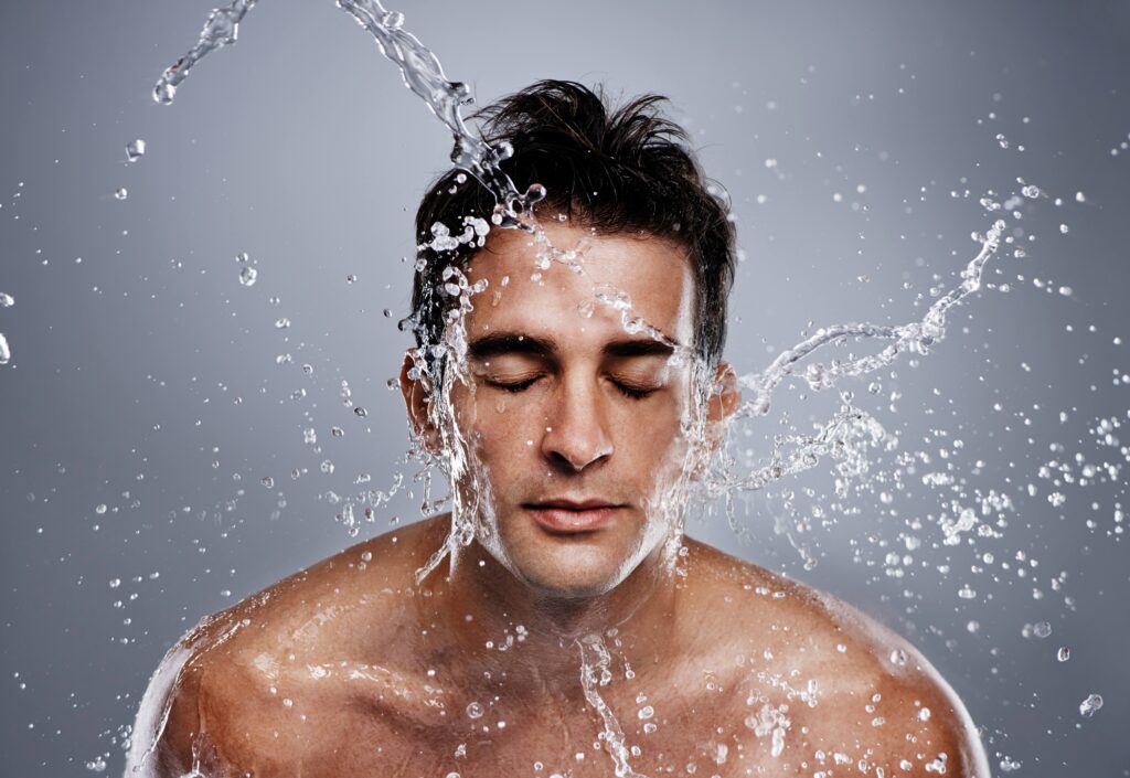 Ein Mann mit nassen Haaren, der die Augen geschlossen hat, während Wasser auf sein Gesicht spritzt. Der Hintergrund ist grau, und das Bild vermittelt ein Gefühl von Frische und Reinheit