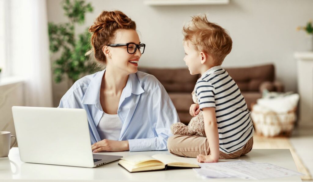 Mutter arbeitet am Laptop, während ihr Kind auf dem Tisch sitzt – Unterstützung durch Familienservice für berufstätige Eltern.