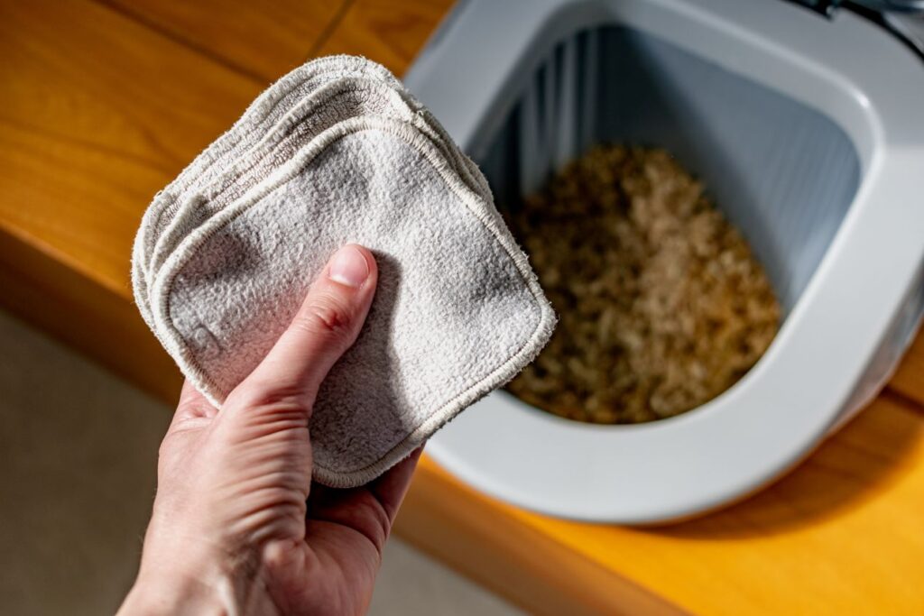 Hand hält waschbare Tücher vor einer geöffneten Trockentrenntoilette – nachhaltige und umweltfreundliche Hygiene.