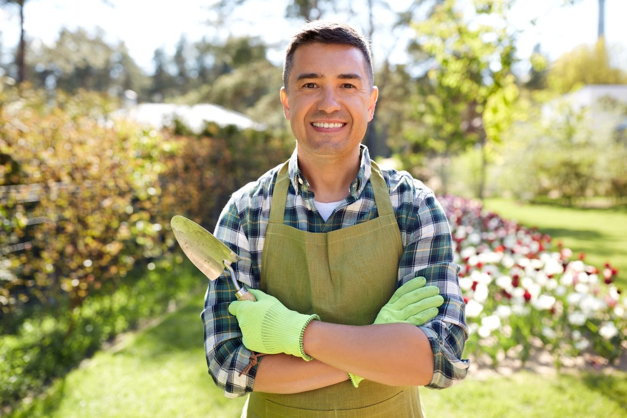 Gärtner mit Schürze und Handschuhen hält eine Schaufel und steht in einem blühenden Garten – wertvolle Tipps zur Hochbeet-Gestaltung.