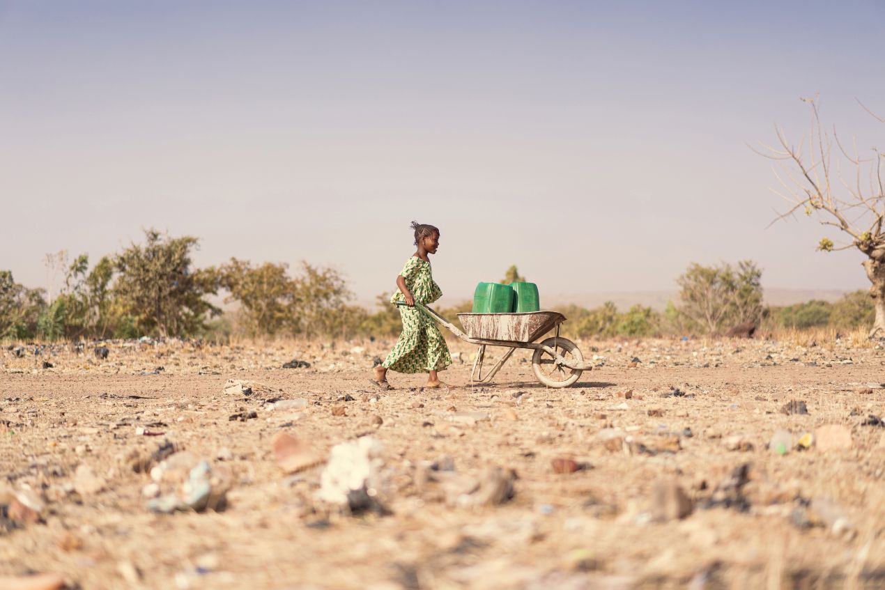 Nothilfe Sudan: Mädchen schiebt Schubkarre mit Wasserkanistern durch trockenes Gebiet ohne Infrastruktur