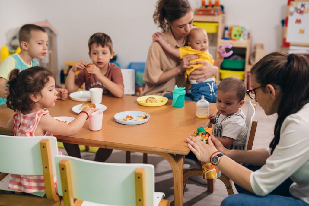 Mehrere Kinder essen gemeinsam an einem Tisch im Kindergarten Schwabing, betreut von zwei Erzieherinnen