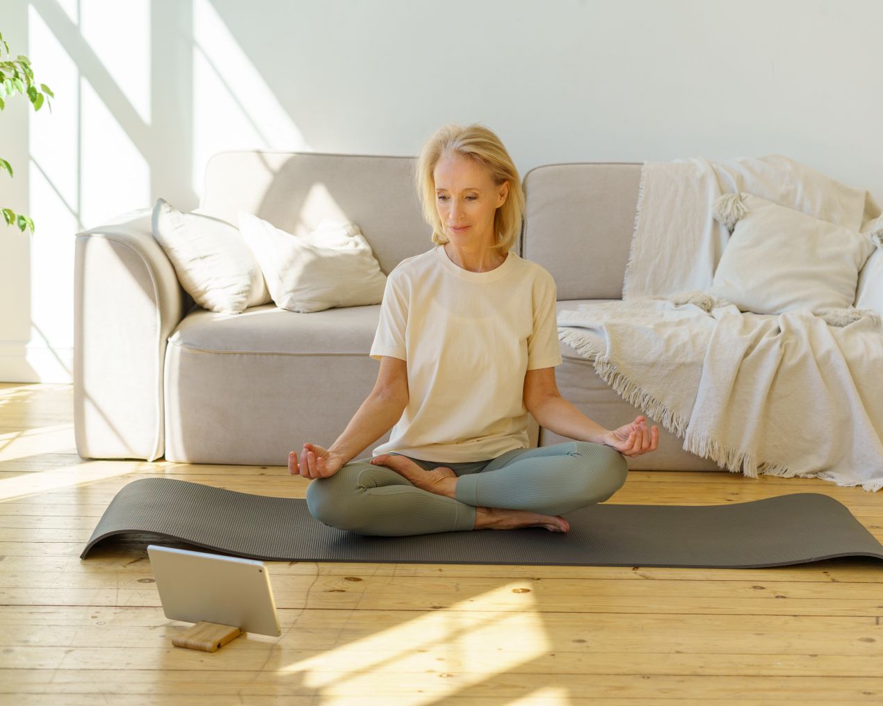 Aeltere Frau sitzt im Schneidersitz auf einer Yogamatte im Wohnzimmer und macht eine gefuehrte Breathwork-Einheit mit einem Tablet